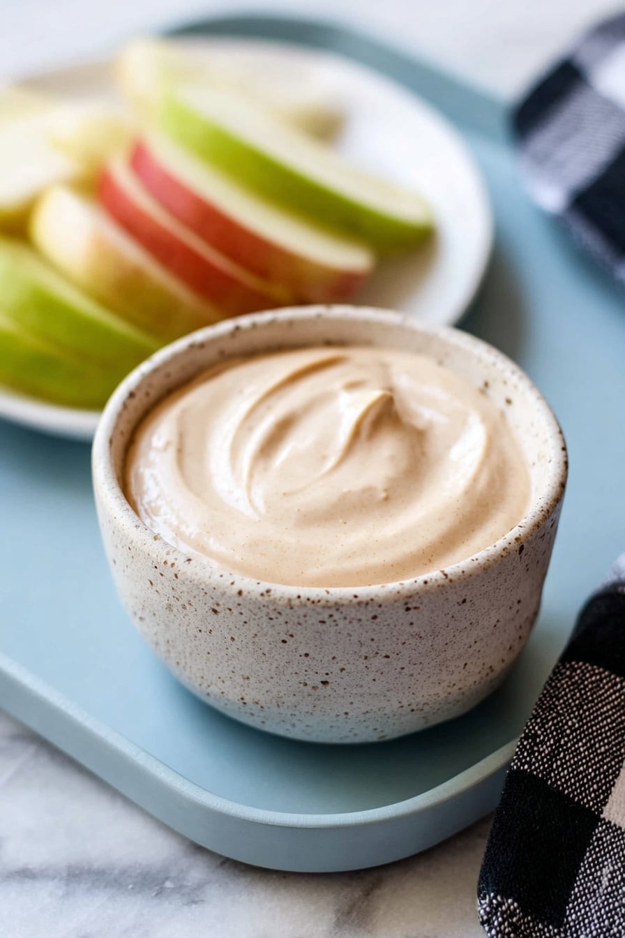 The image shows a small speckled ceramic bowl filled with a smooth, creamy, light beige dip that has soft swirls on the surface. Behind the bowl, there is a white plate with sliced green and red apple wedges, arranged loosely. The bowl and plate are placed on a light blue tray, which rests on a white marbled surface. A black and white checkered cloth is seen partly to the right side. Photo taken with an iphone --ar 2:3 --v 7 - Caramel Apple Dip, easy caramel apple dip, best caramel apple dip recipe, quick apple dip, creamy caramel apple dip