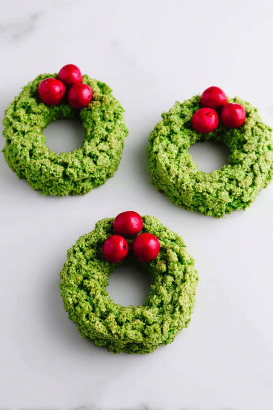 Three small green wreath-shaped treats lie on a white marbled surface. Each wreath is made of a textured, uneven green layer that looks crunchy and dense, shaped in a round ring with a hollow center. On each wreath, there are three glossy red round berries placed together on one side, adding a bright contrast to the green. The arrangement is simple and neat, with the wreaths evenly spaced in a row. photo taken with an iphone --ar 2:3 --v 7 - Easy Cornflake Wreaths, Christmas snack ideas, holiday treats for kids, festive no-bake desserts, quick Christmas cookies
