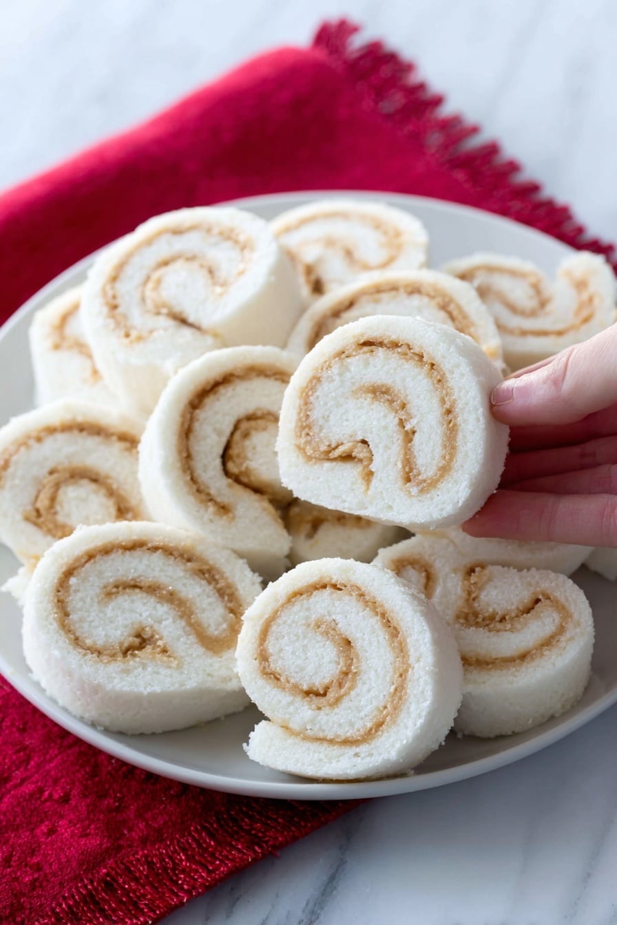 The image shows a white plate filled with soft, spiral-shaped rolls arranged closely together. Each roll has a smooth white outer layer with swirled light tan filling inside, creating a clear contrast. The rolls look fluffy and neatly sliced, with the spirals visible on the sides. The plate is placed on a bright red cloth, and one roll is in the foreground, slightly separated. The background and surface have a white marbled texture. A woman's hand is holding one roll in the top right corner. photo taken with an iphone --ar 2:3 --v 7 - Potato Candy Pinwheels with Peanut Butter, potato candy recipe, peanut butter pinwheels, nostalgic candy recipes, easy homemade candy