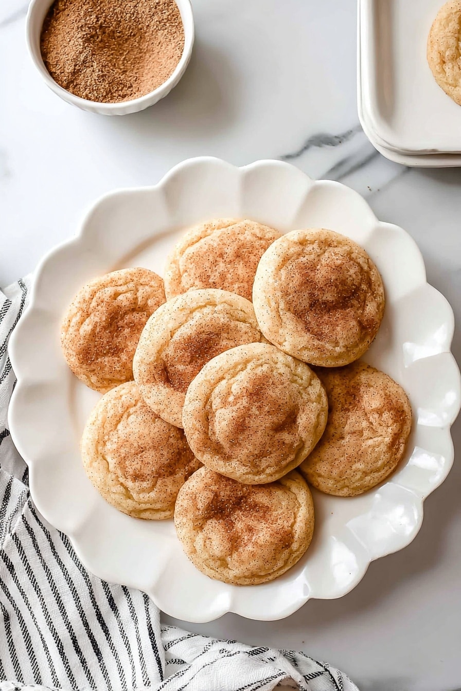 There are seven round snickerdoodle cookies with a light golden brown color and a dusting of cinnamon sugar on top, arranged in a flower shape on a white scalloped plate. The cookies have a soft, slightly wrinkled texture and are placed on a white marbled surface. Near the top left of the plate, there is a small white bowl filled with cinnamon sugar. A white cloth with thin black stripes lays slightly under the plate on the left side. Photo taken with an iphone --ar 2:3 --v 7 - Easy Soft Snickerdoodle Cookies, best snickerdoodle cookies, soft cinnamon sugar cookies, chewy snickerdoodle recipe, quick homemade snickerdoodles