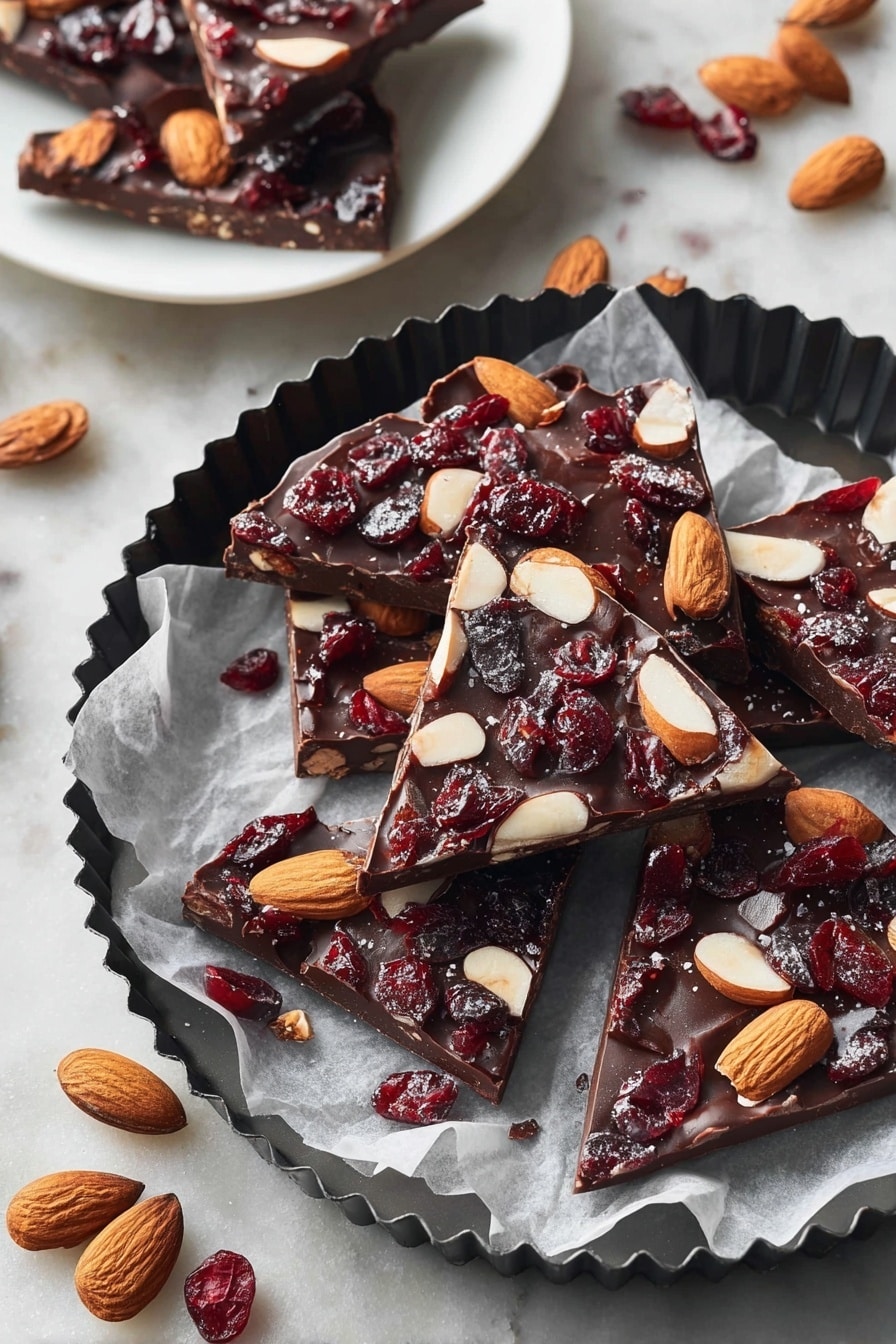 The image shows several pieces of chocolate bark, each with one thick layer of dark brown chocolate topped with whole light brown almonds and dark red dried cranberries scattered evenly. The chocolate pieces are irregular triangles and squares placed on white parchment paper inside a black fluted round pan. Around the pan are whole almonds and dried cranberries on a white marbled textured surface. In the background, a white plate holds three more pieces of chocolate bark, showing the same dark chocolate base with almonds and cranberries. photo taken with an iphone --ar 2:3 --v 7 - Easy Chocolate Bark, chocolate bark with nuts and dried fruit, simple chocolate bark recipe, quick homemade chocolate treat, customizable chocolate bark