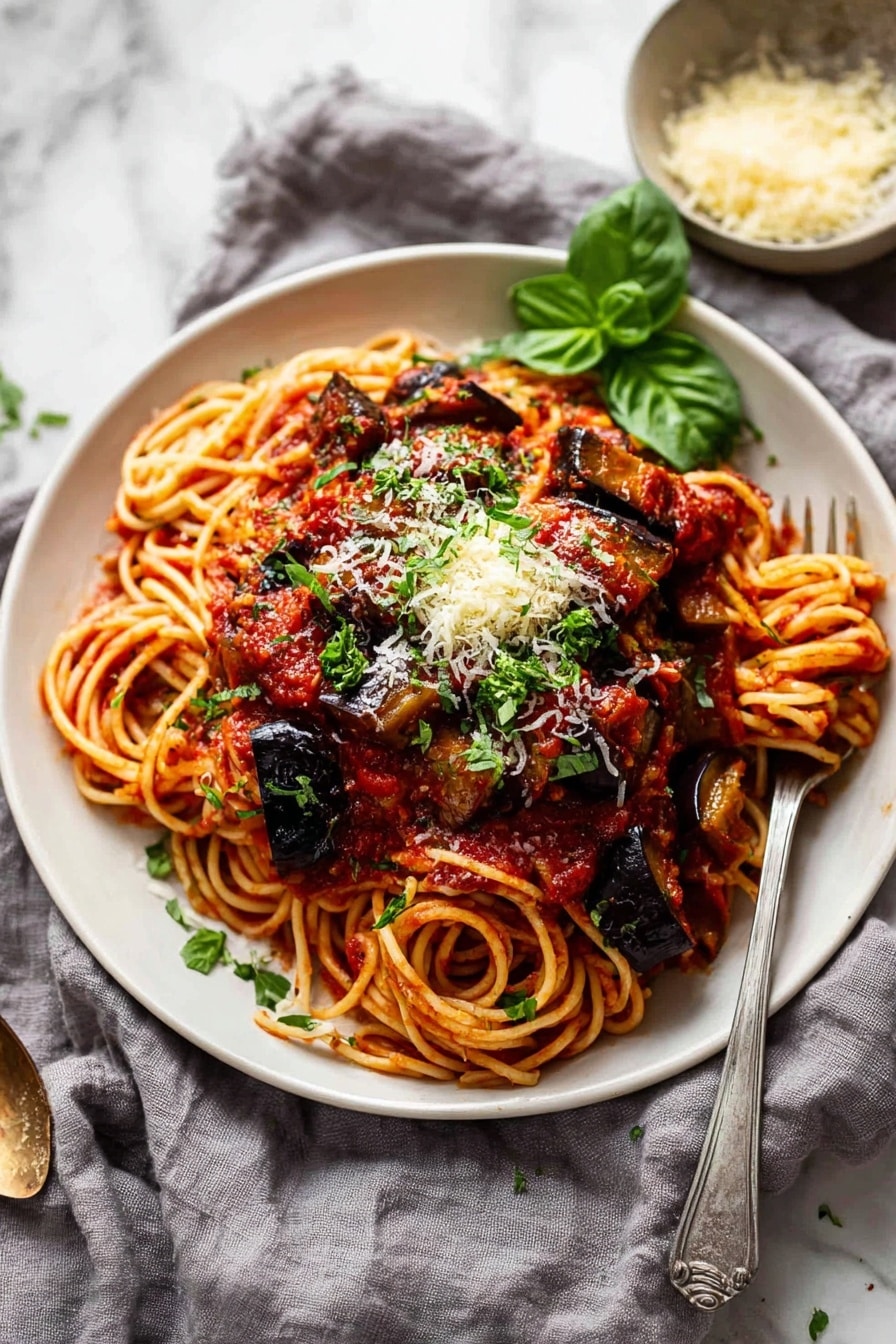 The image shows a close-up of a white bowl filled with spaghetti pasta soaked in rich red tomato sauce, with thick chunks of cooked eggplant scattered on top. The spaghetti strands are tangled at the base and coated evenly with sauce. Large pieces of dark purple eggplant, slightly charred and shiny, sit on the pasta, sprinkled with light yellow grated cheese and small green herb leaves. A silver fork is partly stuck into the pasta, lifting some strands with sauce. The bowl rests on a white marbled surface. Photo taken with an iphone --ar 2:3 --v 7 - Eggplant Pasta alla Norma, Eggplant pasta recipe, Sicilian eggplant pasta, Mediterranean pasta dish, easy Italian eggplant pasta