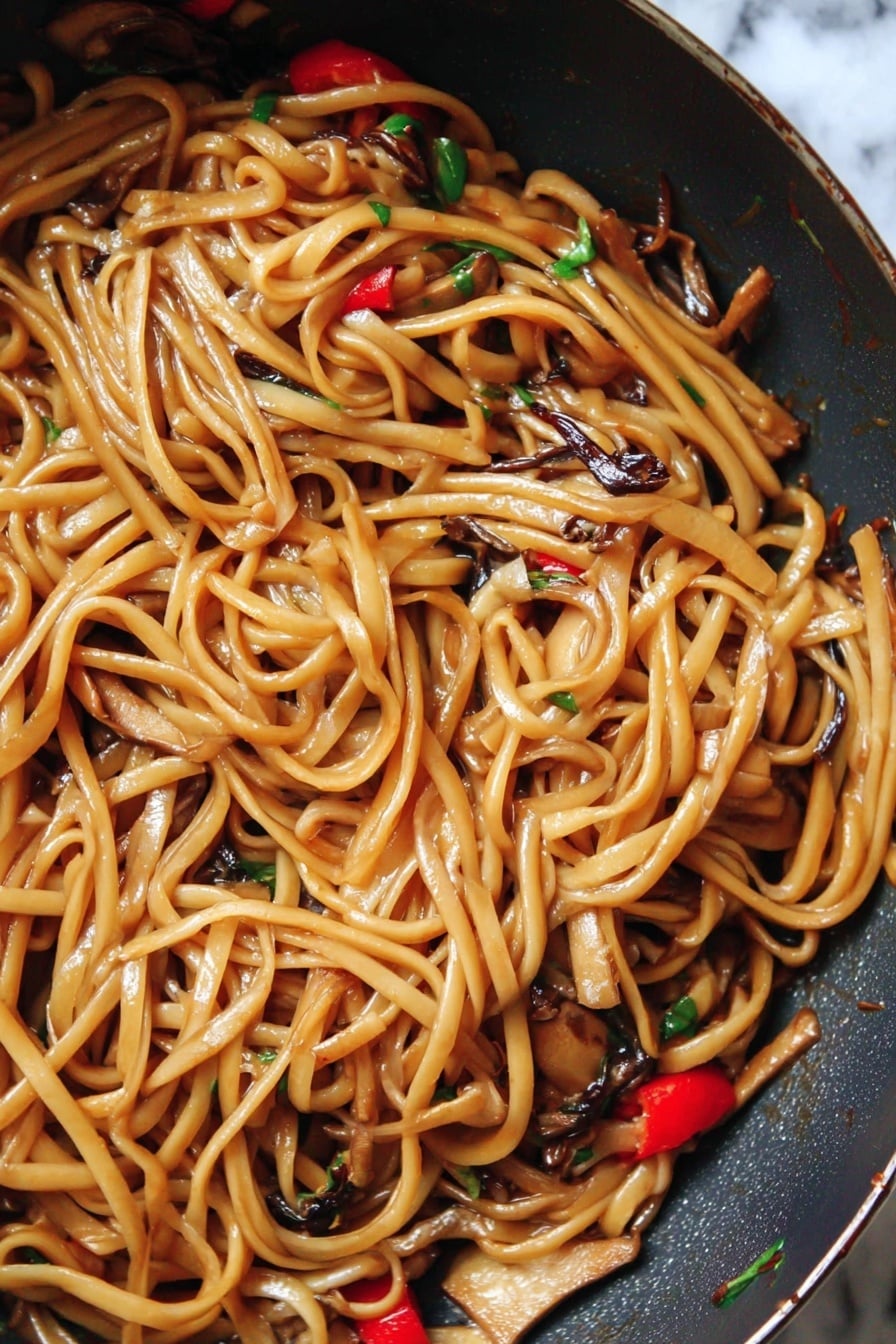 The image shows a close-up of a pan filled with long, shiny noodles coated in a light brown sauce. Mixed among the noodles are small pieces of light tan tofu, dark brown thin mushroom strips, and bright red chili slices, with tiny bits of green herbs scattered throughout. The noodles look soft and glossy, with a slight twist and curl to them, giving a textured, tangled appearance. The pan has a dark surface and the background is changed to a white marbled texture. photo taken with an iphone --ar 2:3 --v 7 - Vegan Kung Pao Pasta with Mushrooms, vegan pasta recipes, Kung Pao pasta recipe, plant-based mushroom pasta, spicy vegan pasta dishes