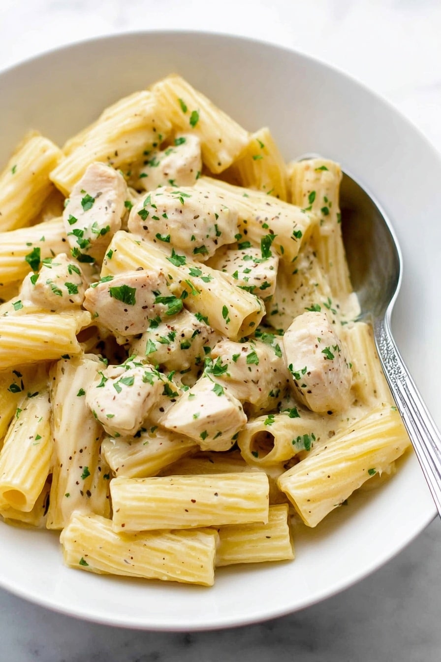 A white bowl filled with creamy rigatoni pasta mixed with chunks of light beige chicken pieces, all coated in a smooth, pale yellow sauce. The pasta is ribbed and tubular, scattered evenly throughout the dish. Small green parsley flakes are sprinkled over the top, adding bright color contrast. A silver spoon rests inside the bowl on the right side, partially under the pasta. The bowl sits on a white marbled surface. Photo taken with an iphone --ar 2:3 --v 7 - Creamy Lemon Chicken Pasta, lemon chicken pasta, creamy pasta recipes, quick chicken pasta, lemon chicken dinner