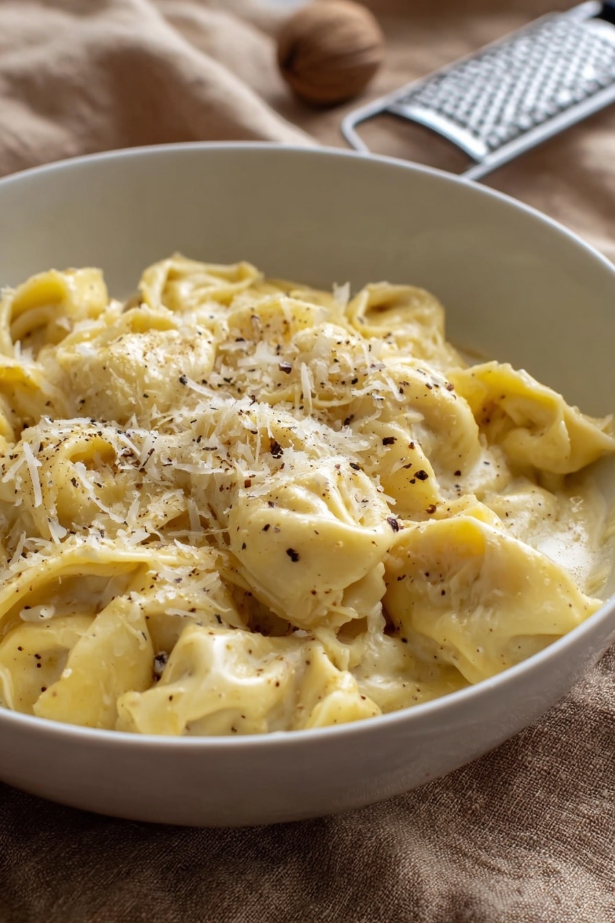 A white bowl filled with about two layers of soft, folded tortellini pasta in a creamy, pale yellow sauce. The pasta is sprinkled with finely grated white cheese and small black pepper flakes, mostly concentrated on the top center. The bowl rests on a tan textured cloth, with a metal grater and a whole nutmeg blurred in the background. The lighting is natural and soft, showing the smooth creamy texture of the sauce and the delicate folds of the tortellini photo taken with an iphone --ar 2:3 --v 7 - Creamy Cheese Tortellini, cheese tortellini recipe, easy pasta dinner, quick creamy pasta, comfort food recipes