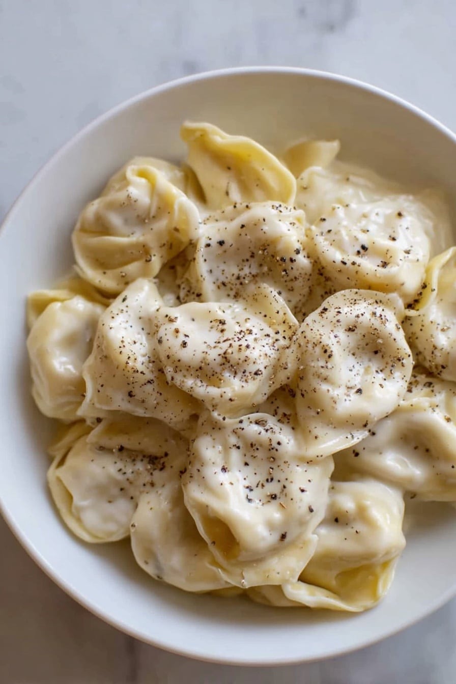 A white bowl filled with a single layer of tortellini pasta, covered evenly with a smooth, creamy white sauce that has a slightly shiny texture. The tortellini are pale yellow with soft folds, and black pepper is sprinkled generously on top, adding small dark specks scattered over the sauce. The bowl is placed on a white marbled surface, and the light highlights the soft texture of both the pasta and sauce photo taken with an iphone --ar 2:3 --v 7 - Creamy Cheese Tortellini, cheese tortellini recipe, easy pasta dinner, quick creamy pasta, comfort food recipes