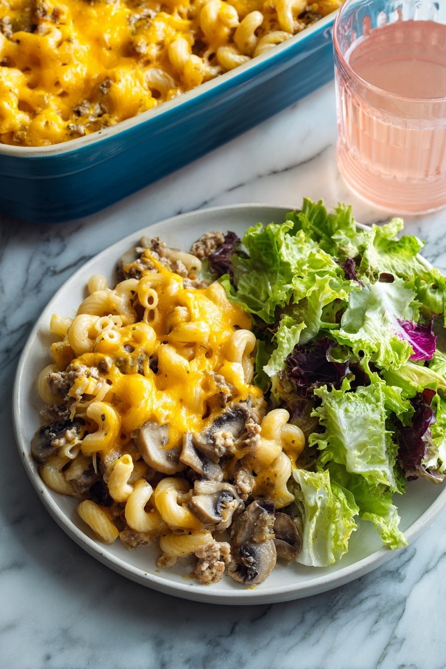 The image shows a white round plate with two main parts: on the left, a layer of baked macaroni mixed with ground meat and sliced mushrooms, all topped with melted bright yellow-orange cheese that looks gooey and soft, and on the right, a fresh green salad made of different lettuce leaves with a mix of light and dark green colors and some purple edges. The plate is set on a white marbled surface, and in the background, there is a blue baking dish filled with more of the macaroni bake and a clear glass of light pink drink. Photo taken with an iphone --ar 2:3 --v 7 - Beef Mushroom Johnny Marzetti Casserole, hearty casserole with beef and mushrooms, easy comfort food recipe, cheesy beef mushroom bake, classic American casserole