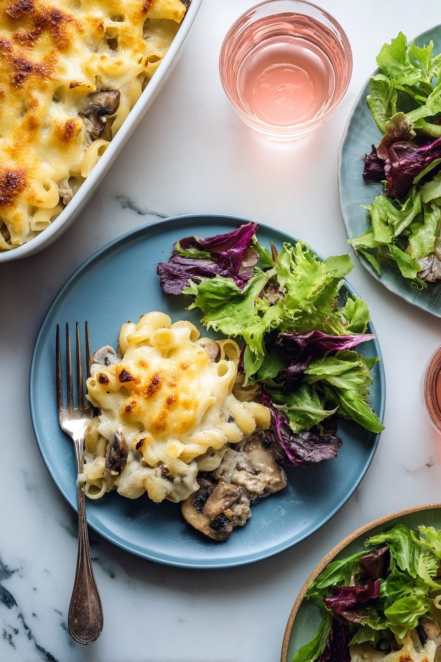 A blue plate holds a serving of baked macaroni and cheese with visible pieces of mushrooms and ground meat under a layer of melted golden-yellow cheese on the left side; on the right side of the plate, there is a fresh green salad made of mixed leafy greens with different shades of green and purple. A silver fork rests on the plate's left edge underneath the macaroni. In the background, a white baking dish filled with more macaroni and cheese sits on a white marbled surface along with a small clear glass of light pink drink. Another plate with leafy greens and another glass of pink drink are partially visible on the right side. photo taken with an iphone --ar 2:3 --v 7 - Beef Mushroom Johnny Marzetti Casserole, hearty casserole with beef and mushrooms, easy comfort food recipe, cheesy beef mushroom bake, classic American casserole
