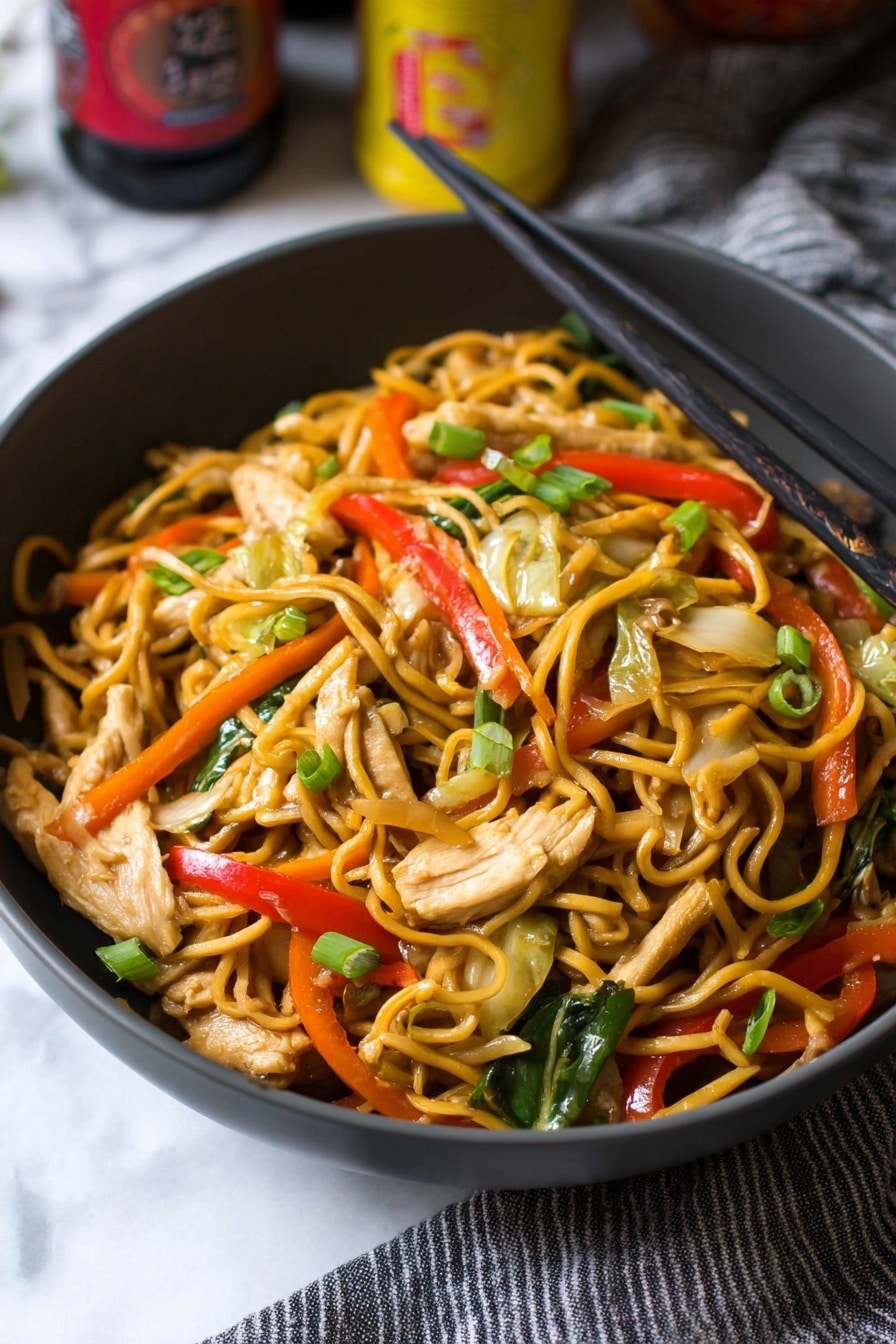 A close-up view of a dark gray bowl filled with stir-fried noodles mixed with thin strips of light beige chicken pieces, bright red bell pepper slices, green scallion pieces, thin orange carrot strips, light green cabbage, and sliced beige mushrooms. Black chopsticks rest on the right edge of the bowl, while the bowl sits on a white marbled surface with part of a gray striped cloth to the right. The background includes some blurred bottles with yellow and red labels. photo taken with an iphone --ar 2:3 --v 7 - Chicken Yakisoba Stir-Fry, Chinese stir-fry recipes, quick Asian noodle dishes, homemade yakisoba, veggie stir-fry recipes