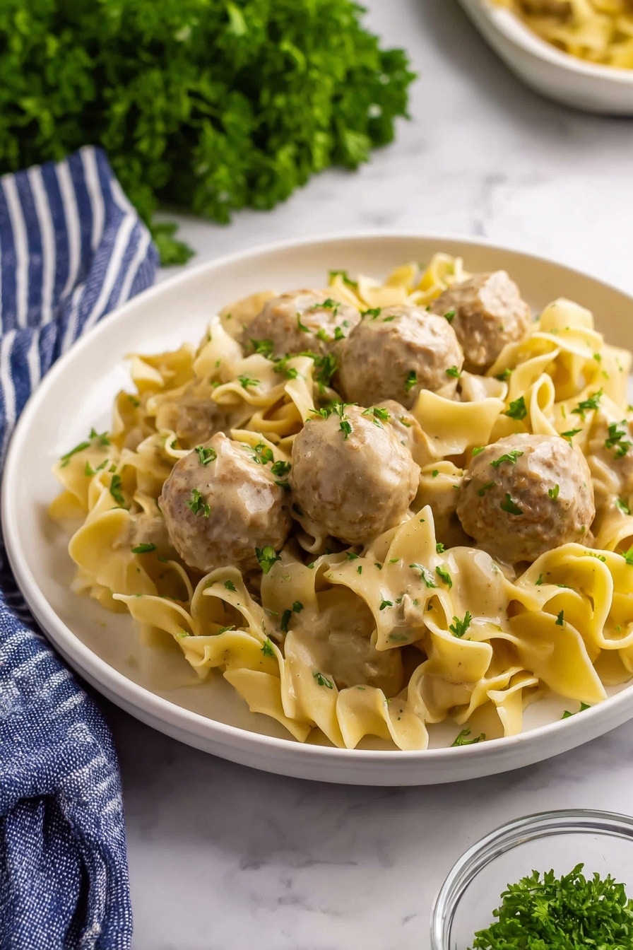 A white plate holds a serving of wide, curly egg noodles mixed with round meatballs covered in a creamy light brown sauce. The noodles are soft and slightly twisted, with flecks of green herbs sprinkled on top. The meatballs are light brown, smooth, and sit evenly spaced among the noodles. Bright green parsley leaves are scattered across the dish, adding color contrast. The scene is set on a white marbled surface with a bunch of fresh parsley blurred in the background, a blue and white striped cloth partially visible at the bottom left, and a small glass bowl with chopped herbs near the plate. Photo taken with an iphone --ar 2:3 --v 7 - Creamy Swedish Meatball Noodle Skillet, Swedish meatball skillet recipe, comforting Swedish-inspired dinner, easy one-pan meatball meal, quick creamy meatball dinner