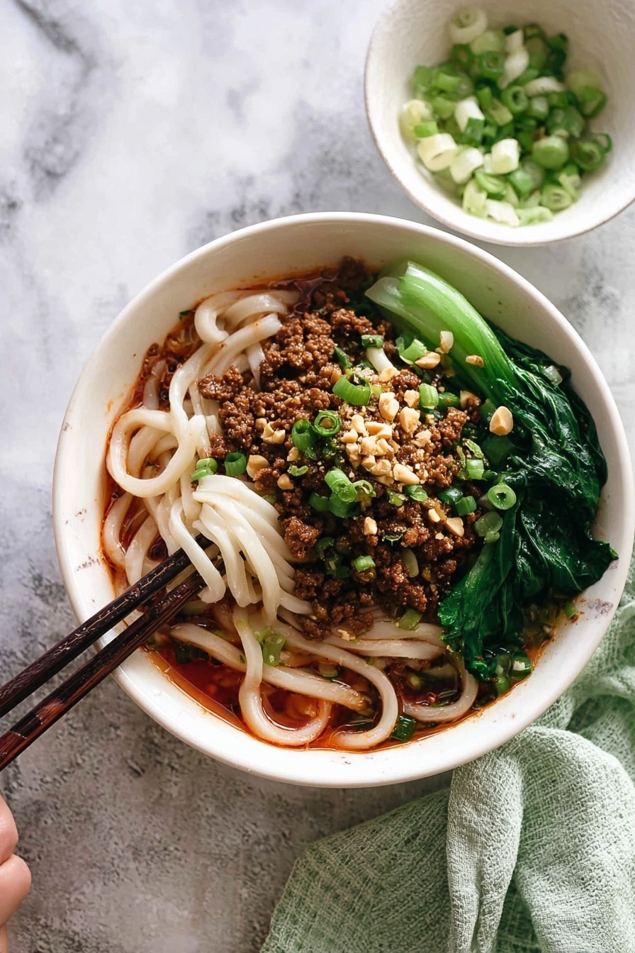 The image shows a white bowl filled with three main layers: at the bottom, thick white noodles soaked in a reddish-brown sauce, in the middle, crumbled cooked meat with chopped green onions and crushed peanuts sprinkled on top, and on the side, bright green leafy vegetables placed neatly. A pair of dark wooden chopsticks is picking up some noodles from the bowl. Next to the bowl, there is a small white bowl with sliced green onions, and a light green towel is folded in the background. The whole scene is set on a white marbled surface. photo taken with an iphone --ar 2:3 --v 7 - Spicy Dan Dan Noodles with Pork, Sichuan Dan Dan Noodles, Homemade Dan Dan Noodles, Pork and Chili Oil Noodles, Authentic Sichuan Noodles