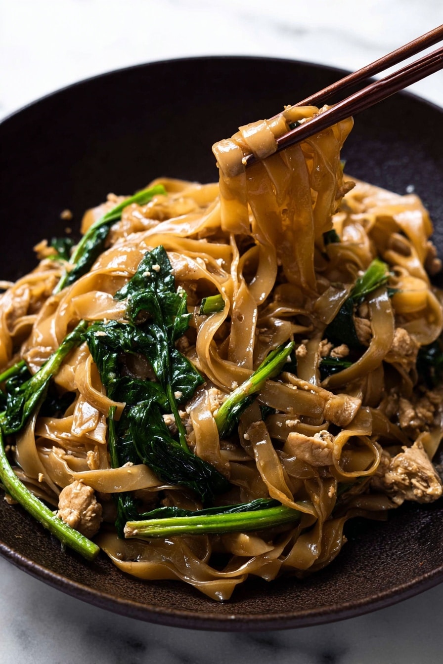 This image shows a close-up of stir-fried noodles in a dark pan. The dish has wide, flat noodles that are light brown and shiny from sauce, mixed with bright green leafy vegetables and light brown pieces of cooked chicken. The noodles and vegetables are tangled together, with the noodles forming the main layer in the center and the green vegetables scattered around. A wooden spatula is seen gently holding some of the noodles from the top right side. The background is a white marbled texture. photo taken with an iphone --ar 2:3 --v 7 - Authentic Thai Pad See Ew with Chicken, Thai noodle stir-fry recipe, easy Thai chicken stir-fry, homemade Pad See Ew, Thai street food at home