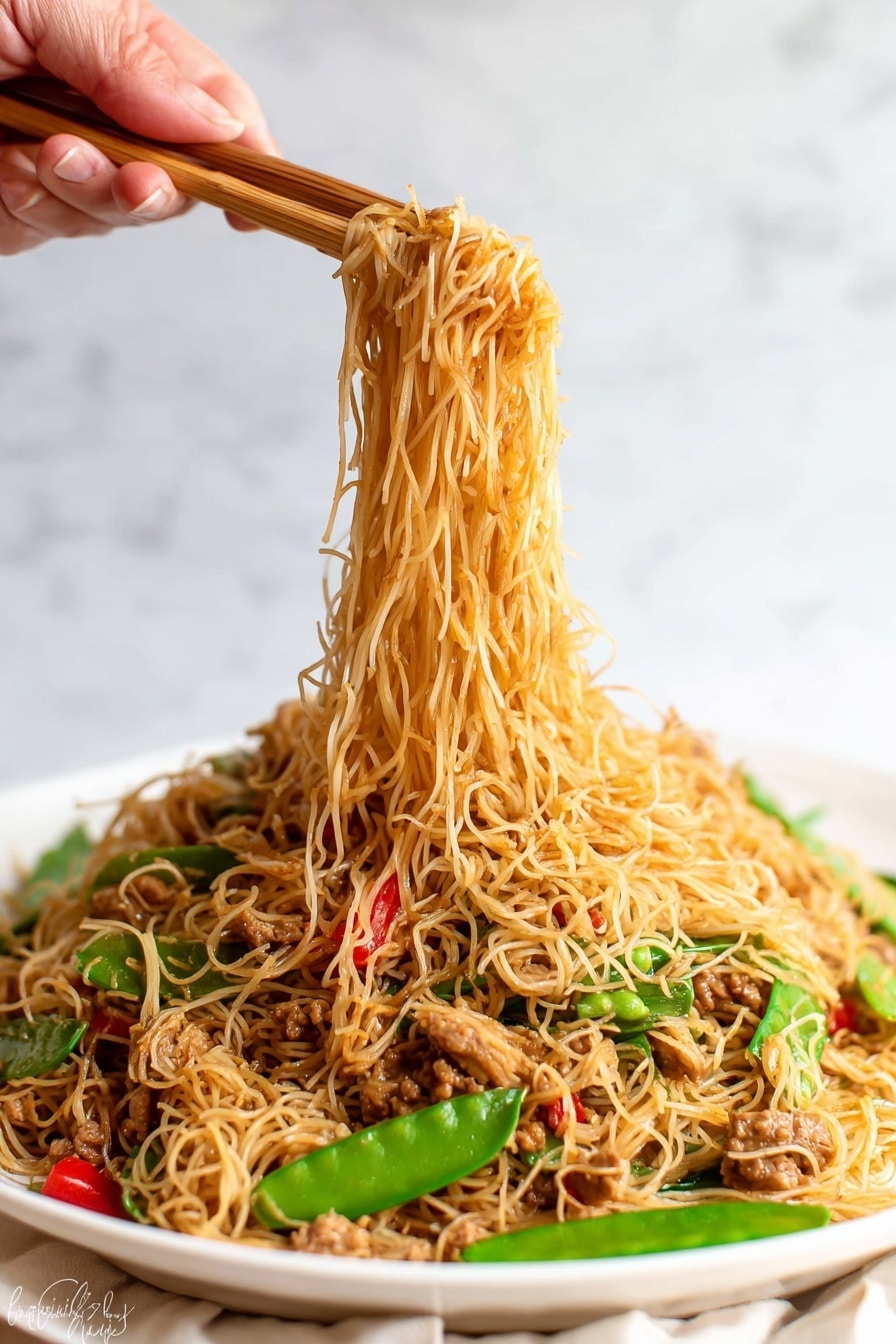 The image shows a white plate full of thin noodles mixed with small pieces of cooked meat, green snow peas, and red bell pepper slices. The noodles are light brown and soft, forming the top and main layer, while the vegetables and meat are scattered throughout, adding pops of green and red colors. A woman's hand is holding wooden chopsticks lifting a portion of the noodles above the plate, with the noodles hanging down in thin strands. The background has a white marbled texture. photo taken with an iphone --ar 2:3 --v 7 - Singapore Curry Noodles Chinese stir-fry, easy curry noodle dish, quick Asian noodle recipe, flavorful curry noodle dinner, weeknight Asian meal