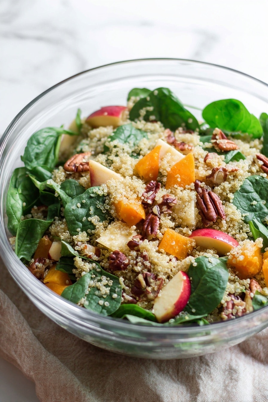 A clear glass bowl holds a salad with three main layers: a bottom layer of light beige quinoa grains scattered throughout, a middle layer of bright green fresh spinach leaves placed on top and mixed in, and a top layer consisting of medium-sized chunks of orange squash, small cubes of red and yellow apple pieces, light beige chickpeas, and whole brown pecan nuts arranged evenly around the salad. Next to the bowl is a small white bowl filled with pecans. The setting includes two vintage silver salad servers placed on a white and red striped cloth, all set on a white marbled texture surface. photo taken with an iphone --ar 2:3 --v 7 - Roasted Butternut Squash Quinoa Salad, healthy butternut squash salad, autumn quinoa salad, easy roasted squash salad, gluten-free quinoa salad