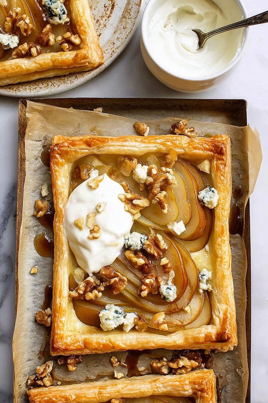 The image shows a golden brown puff pastry tart with a raised edge surrounding soft, thinly sliced caramelized pears arranged in an overlapping pattern. On top of the pears, there are small chunks of blue cheese and scattered walnut pieces adding texture and color contrast. A dollop of creamy white yogurt or cream is placed on one corner of the tart. The tart sits on parchment paper which lines a baking tray with some walnut pieces and cheese crumbs around it. Nearby, a small white ceramic bowl with a spoon inside contains more white cream. The background is a white marbled surface. photo taken with an iphone --ar 2:3 --v 7 - Caramelized Pear and Blue Cheese Tart, pear and blue cheese tart, caramelized pear tart, blue cheese pastry tart, easy savory tart