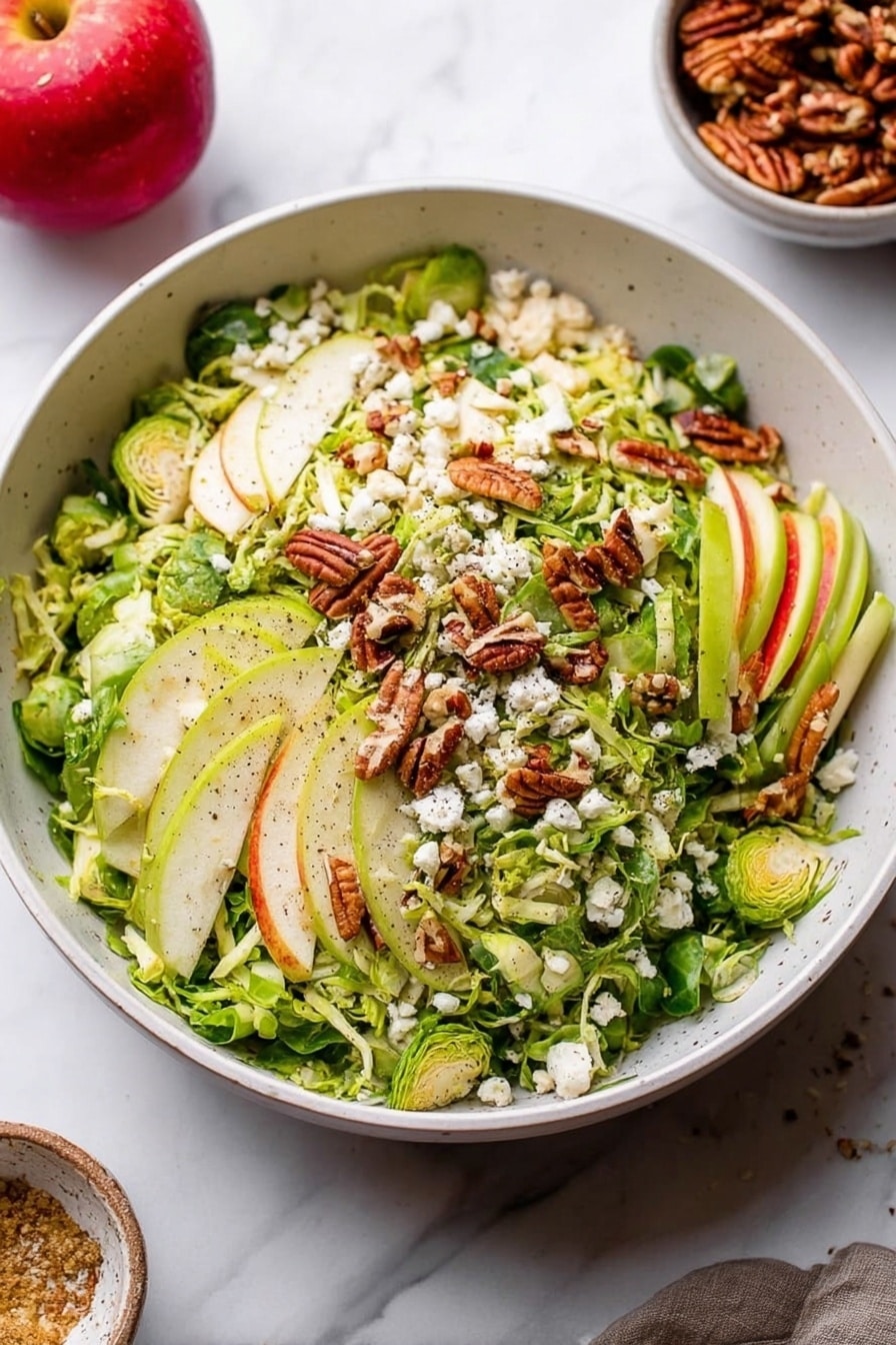 A large white bowl filled with a fresh salad featuring several layers: the base layer is thinly sliced green Brussels sprouts, topped with light green and white strips of apple. Scattered on top are golden-brown pecans and small white crumbles of cheese. The ingredients have a fresh, slightly rough texture with visible black pepper sprinkled over. The bowl sits on a white marbled surface, and in the background, a red apple and a small bowl of pecans are visible. Photo taken with an iphone --ar 2:3 --v 7 - Fall Brussels Sprout Salad with Apples and Blue Cheese, Brussels Sprouts Salad, Autumn Side Salad, Easy Fall Recipes, Healthy Fall Salad