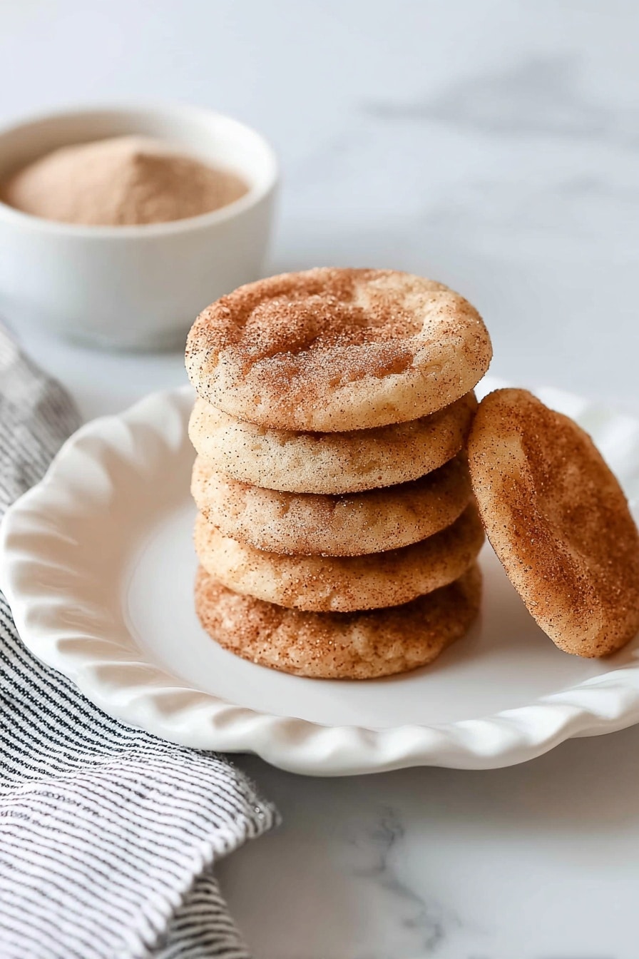 A stack of eight round, light brown cookies with a dusting of cinnamon sugar on top sits in the center of a white plate with scalloped edges. One cookie lies flat behind the stack, showing its darker cinnamon spots. The white plate is placed on a white marbled surface with a folded white and black striped cloth partially visible on the left. In the top left corner, a small white bowl filled with a light brown powder is slightly out of focus. photo taken with an iphone --ar 2:3 --v 7 - Easy Soft Snickerdoodle Cookies, best snickerdoodle cookies, soft cinnamon sugar cookies, chewy snickerdoodle recipe, quick homemade snickerdoodles