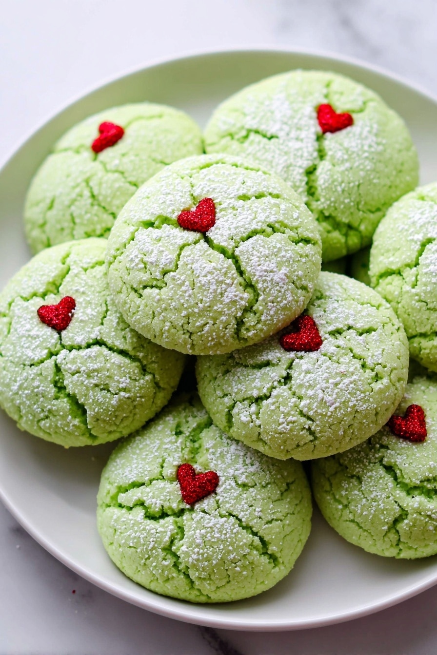 The image shows a white plate filled with round green cookies that have a cracked texture on top. Each cookie has a light dusting of white powder and a tiny red heart decoration placed slightly off-center on the top surface. The cookies are stacked closely together on the plate, which sits on a white marbled background. Photo taken with an iphone --ar 2:3 --v 7 - Grinch Cookies with Peppermint and Green Food Coloring, festive holiday cookies, Christmas cookie ideas, peppermint cookies recipe, easy holiday treats