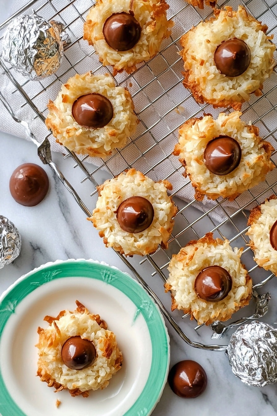The image shows seven round coconut cookies with a light golden-brown crispy outside and white, shredded coconut texture in the middle layer. Each cookie has a single smooth milk chocolate drop placed in the center on top. The cookies are arranged on a silver cooling rack that rests on a white rectangular marble slab. Around the rack, there are several extra chocolate drops scattered on the white marbled surface. A woman's hand is holding one cookie on a white plate with a green rim, and a few silver-wrapped chocolate candies are placed nearby. The overall setting is bright with a white marbled background. photo taken with an iphone --ar 2:3 --v 7 - Coconut Macaroon Blossoms with Hershey Kisses, coconut cookie recipe, chewy coconut treats, gluten-free coconut cookies, chocolate coconut cookies