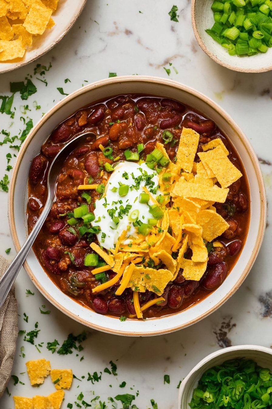 The image shows a white bowl filled with thick chili, dark brown with visible kidney beans and small green herbs mixed in. On top, there is a dollop of white sour cream at the center, surrounded by thin yellow shredded cheese and curved, crunchy-looking yellow corn chips. Chopped green onions and finely chopped green herbs are sprinkled over the sour cream and cheese. A silver spoon rests inside the bowl on the left side. The bowl is placed on a white marbled surface with scattered green herbs and a small white bowl with extra chopped green onions nearby. photo taken with an iphone --ar 2:3 --v 7 - Easy Beef Chili, hearty beef chili, quick chili recipe, easy dinner ideas, beef chili dinner