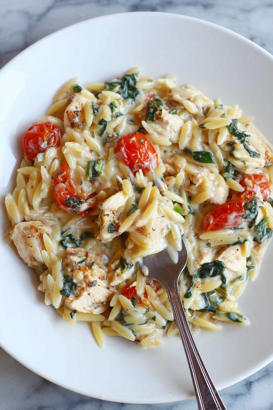 A white round plate holds a creamy pasta dish composed of orzo pasta mixed with small chunks of light brown cooked chicken pieces and bright red cherry tomatoes. There are dark green leafy bits, possibly spinach or herbs, spread evenly throughout the dish. The sauce looks smooth and creamy, coating the pasta and ingredients in a soft white layer. A silver fork is placed inside the pasta near the bottom center of the plate. The plate sits on a white marbled surface photo taken with an iphone --ar 2:3 --v 7 - Creamy Chicken Orzo Bake, Creamy Chicken Orzo, Chicken Orzo casserole, baked chicken and orzo, easy creamy chicken bake