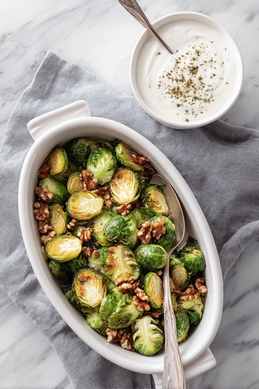 A white oval dish filled with two layers of roasted Brussels sprouts that are deep green with golden brown edges, topped with scattered light brown walnut halves. A silver spoon rests inside the dish, partially submerged in the vegetables. The dish sits on a gray cloth napkin with a white marbled background. To the right, there is a small white bowl filled with white creamy sauce topped with a sprinkling of brown herbs, with a white spoon inside. Photo taken with an iphone --ar 2:3 --v 7 - Roasted Brussels Sprouts with Walnuts and Pomegranate, Brussels Sprouts with Walnuts and Pomegranate, Pomegranate Brussels Sprouts Side Dish, Walnut and Pomegranate Brussels Sprouts, Healthy Brussels Sprouts with Pomegranate