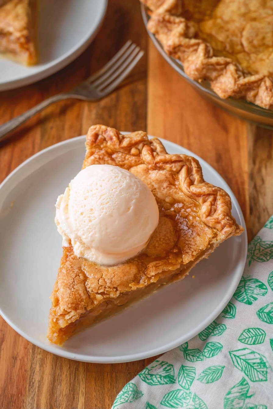 A slice of golden brown pie with a textured, bubbly top crust sits on a white plate. The crust edge is thick and crimped with a wavy pattern, showing a darker toasted color. On top of the pie slice, there is a smooth, creamy scoop of vanilla ice cream with a pale off-white color. Around the plate, the surface is a wooden tabletop with warm tones, and a metal fork lies nearby. Part of the whole pie with a similar bubbly crust and wavy edges is seen in a clear glass pie dish to the upper right. In the lower right corner is a fabric napkin with a green leaf pattern on white. photo taken with an iphone --ar 2:3 --v 7 - Easy Pear Pie, pear dessert, easy fruit pie, cinnamon pear pie, quick pear dessert