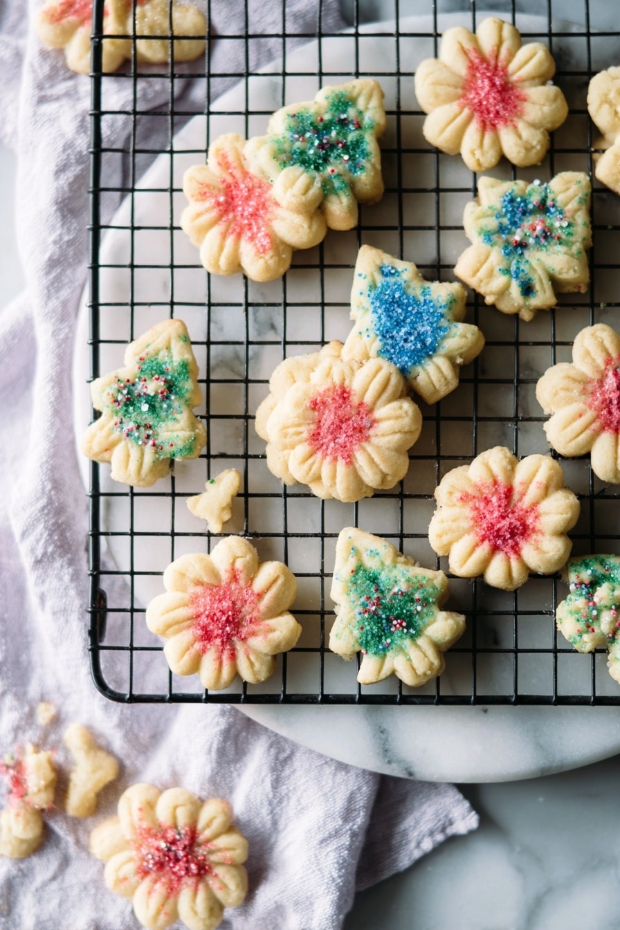 The image shows small, shortbread cookies in two shapes: flower-like rounds and Christmas trees, arranged on a round white marble board and a black wire cooling rack placed on a white marbled surface with a white cloth underneath. The cookies have a light beige color and are decorated with colored sugar sprinkles—red on the round flower-shaped cookies and blue or green on the Christmas tree-shaped cookies. The cookies appear slightly textured with small ridges from the mold, and they are scattered loosely on both the marble board and the wire rack, creating a casual but festive look. Photo taken with an iphone --ar 2:3 --v 7 - Butter Spritz Cookies, Classic Butter Spritz Cookies, Spritz Cookie Recipe, Butter Cookie Shapes, Christmas Spritz Cookies