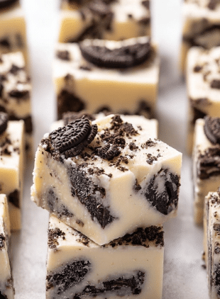 The image shows a close-up of thick square pieces of cookies and cream fudge layered in white creamy base mixed with broken chocolate cookie bits. Each piece has a smooth, pale off-white layer filled with dark brown cookie chunks inside and baked bits scattered on top, creating a textured look with irregular shapes. The fudge pieces are placed closely together on a white marbled surface, highlighting the contrast between the creamy white fudge and dark cookies. Photo taken with an iphone --ar 2:3 --v 7 - White Chocolate Oreo Fudge, easy Oreo fudge, no-bake Oreo dessert, creamy Oreo fudge, Oreo cookie fudge recipe