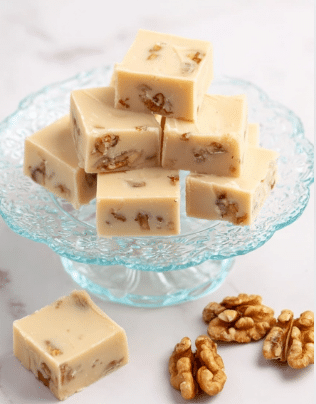 Several square pieces of light beige fudge with small brown nut bits inside are stacked neatly on a white scalloped glass plate with floral patterns. The fudge squares have a smooth and creamy texture, with some pieces resting flat and others leaned against each other. To the right of the plate, a few whole walnut halves are scattered on a white marbled surface. The scene is simple and bright with soft lighting. photo taken with an iphone --ar 2:3 --v 7 - Maple Walnut White Chocolate Fudge, white chocolate fudge with walnuts, easy maple fudge, no-bake holiday treats, creamy walnut fudge