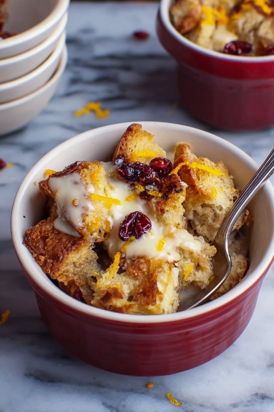 A small white bowl filled with several layers of toasted bread chunks that are golden brown and crispy on the edges. The bread pieces are mixed with creamy, slightly melted white sauce or cream that sits on top and around the bread. There are dark red dried berries scattered on top and some thin orange zest strips add a pop of color. A silver spoon is inserted inside the bowl on the right side. The bowl is placed on a white marbled surface with a small stack of empty white bowls in the background. photo taken with an iphone --ar 2:3 --v 7 - Cranberry Bread Pudding with Toffee Bits, festive holiday bread pudding, easy cranberry dessert, warm cinnamon bread pudding, holiday crowd-pleasing desserts