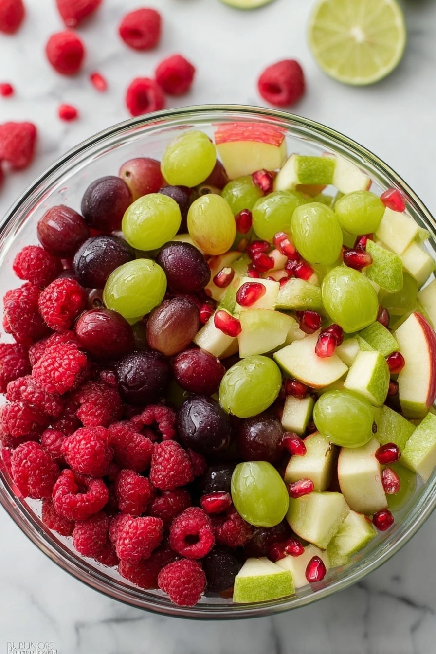 A clear glass bowl filled with a colorful fruit salad sits on a white marbled surface. The salad has multiple layers made up of red raspberries, dark purple grapes, light green grapes, small pieces of green kiwi with black seeds, light yellow apple slices with a red skin edge, and shiny red pomegranate seeds scattered on top. The fruits create a mix of vibrant reds, purples, greens, and yellows with smooth and textured surfaces. Photo taken with an iphone --ar 2:3 --v 7 - Festive Christmas Fruit Salad, Christmas fruit salad, holiday fruit salad, easy Christmas salad, colorful holiday recipe