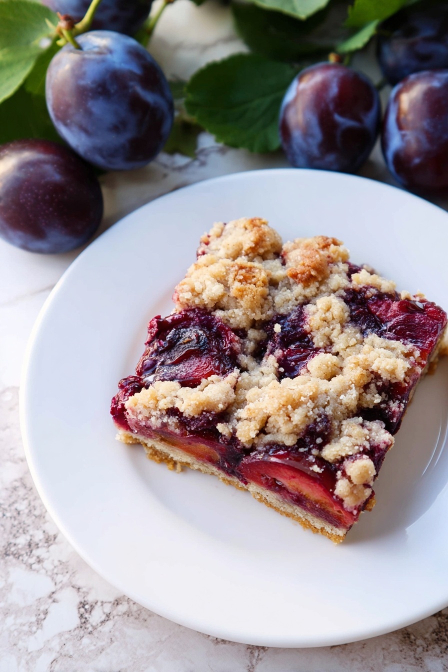 A square slice of a fruit crumble bar sits on a white plate. The bottom layer is a light golden crust, topped with a thick layer of dark red and purple cooked plums showing a soft texture. The top layer is a crumbly streusel with uneven pieces of pale beige and light brown. The plate is placed on a white marbled surface, next to fresh whole dark purple plums with green leaves. Photo taken with an iphone --ar 2:3 --v 7 - German Plum Cake with Yeast Crust, authentic German plum cake, traditional German fruit cake, Swabian plum tart, yeast dough plum dessert