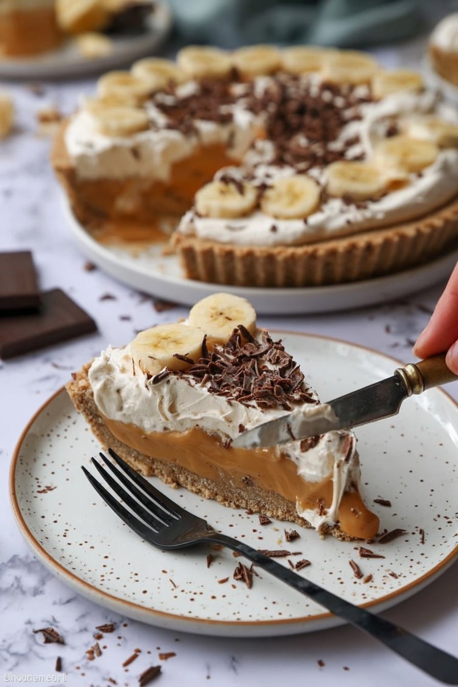 A slice of pie sits on a white plate with brown speckles, showing three layers: a crunchy light brown crust, a smooth caramel-colored filling with banana slices, and a creamy white topping sprinkled with dark chocolate curls. Behind it, the rest of the pie with the same layers is on a white plate, with one slice missing. A woman’s hand holds a silver knife with some pie filling and cream on it. A black fork rests on the plate near the slice. The background is a white marbled surface with some chocolate shavings scattered around. Photo taken with an iphone --ar 2:3 --v 7 - Easy No-Bake Banoffee Pie, no-bake dessert recipes, simple banana pie, quick indulgent desserts, no oven banana pie