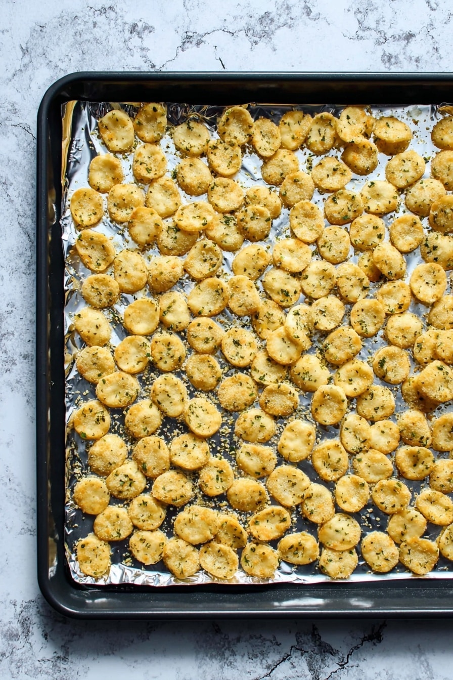 A black baking tray lined with shiny aluminum foil holds a single flat layer of small, round, golden-brown crackers sprinkled with green herbs. The crackers are spread evenly, with slight overlaps in places, showing a crispy and slightly rough texture. The background features a white marbled surface that contrasts with the baking tray. photo taken with an iphone --ar 2:3 --v 7 - Ranch Oyster Crackers Snack, Ranch Oyster Crackers, Easy Snack Recipes, No-Bake Crunchy Snacks, Party Snack Ideas