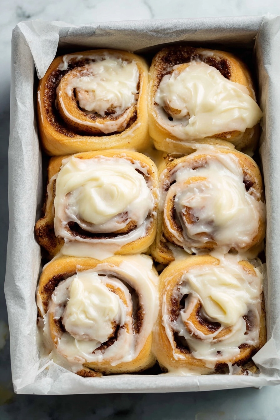 This image shows eight cinnamon rolls in a square baking pan lined with white parchment paper, placed on a white marbled surface. Each roll has two visible layers: the base layer is soft, light golden dough spiraled with a dark brown cinnamon filling, and the top layer is thick, creamy white frosting spread unevenly, melting slightly into the rolls. The rolls are packed closely together, creating a warm, inviting look with some frosting softly dripping between them. Photo taken with an iphone --ar 2:3 --v 7 - Ultimate Fluffy Cinnamon Rolls, homemade cinnamon rolls, soft cinnamon rolls with cream cheese frosting, easy cinnamon roll recipe, melt-in-your-mouth cinnamon rolls