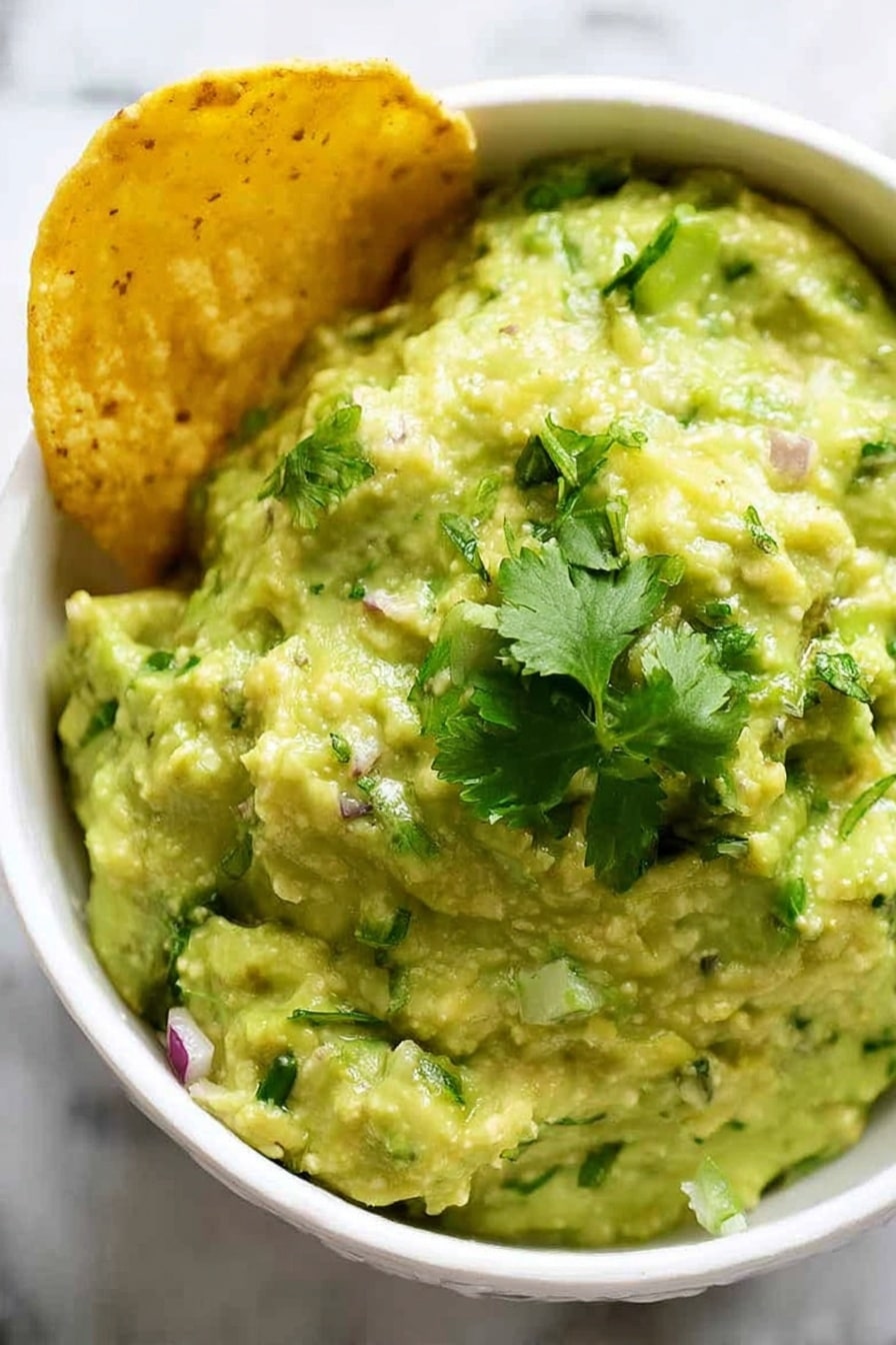 A close-up view of a white bowl filled with creamy, chunky pale green guacamole mixed with small bits of green herbs and red onion, topped with fresh bright green cilantro leaves. On the left side of the bowl, a golden yellow corn chip is partially dipped into the guacamole. The bowl sits on a white marbled surface. photo taken with an iphone --ar 2:3 --v 7 - Quick and Creamy Guacamole, best guacamole dip, easy guacamole recipe, homemade guacamole, party dip ideas