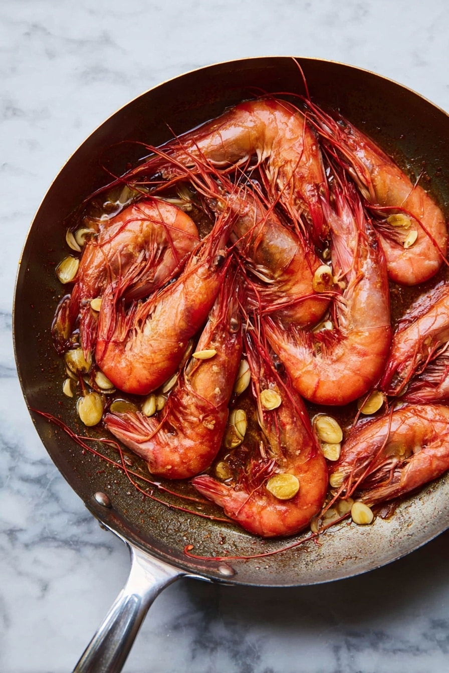 A frying pan filled with cooked whole red prawns layered over scattered golden brown slices of sautéed garlic. The prawns are arranged in a slightly overlapping way, showing their curved, shell-covered bodies, long antennae, and black eyes. The pan has a shiny metal handle extending to the side, and the background is a white marbled surface. photo taken with an iphone --ar 2:3 --v 7 - Garlic Shrimp Skillet, Shrimp skillet recipe, quick garlic shrimp, easy seafood dinner, flavorful shrimp stir-fry