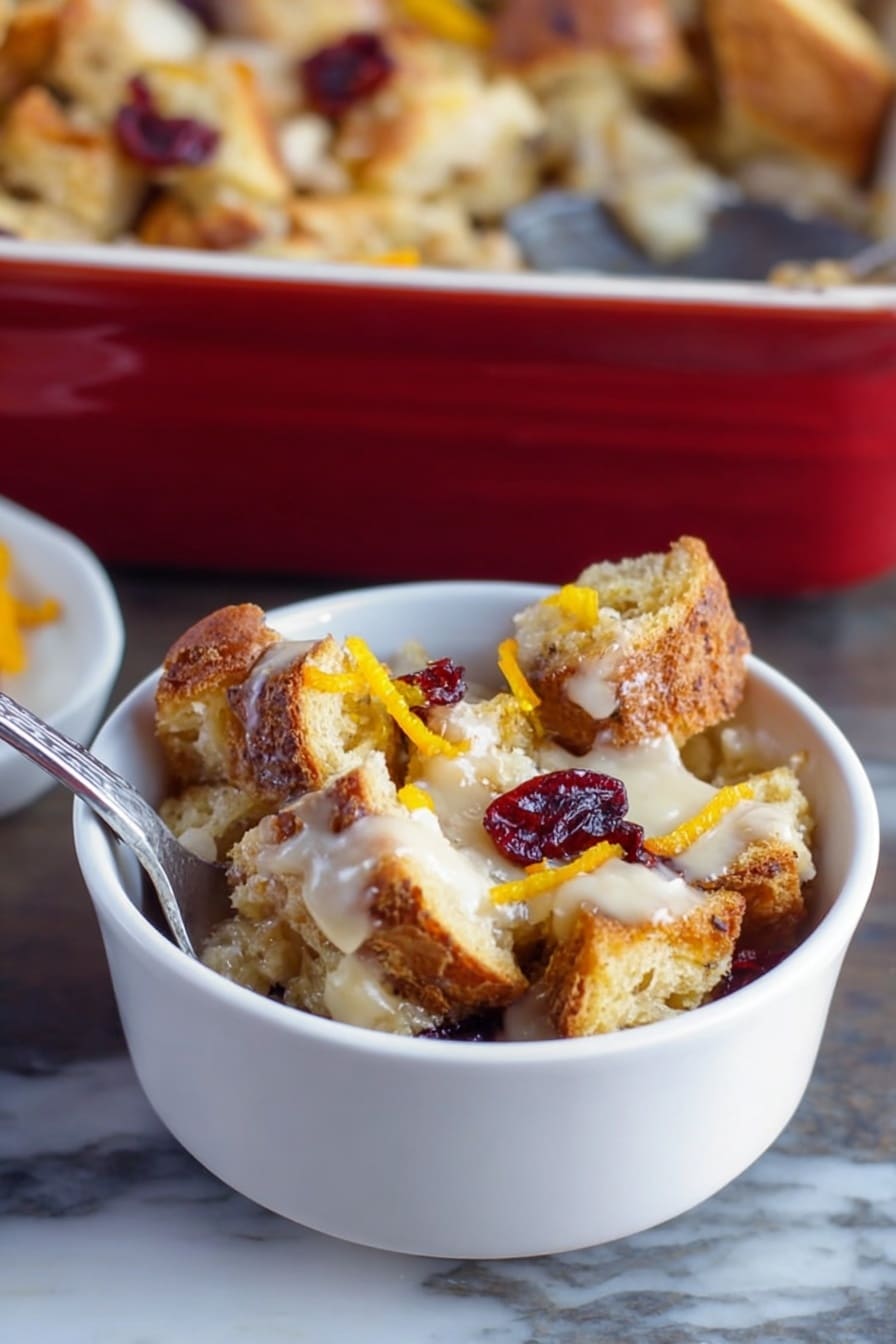 A small white bowl filled with several uneven layers of toasted bread pieces that are golden brown and crispy, mixed with a creamy, light-colored sauce spread unevenly throughout, topped with a few dried red cranberries and thin strips of bright orange zest, placed on a white marbled surface with a silver spoon inside the bowl, a larger baking dish filled with similar bread pudding in the background, photo taken with an iphone --ar 2:3 --v 7 - Cranberry Bread Pudding with Toffee Bits, festive holiday bread pudding, easy cranberry dessert, warm cinnamon bread pudding, holiday crowd-pleasing desserts