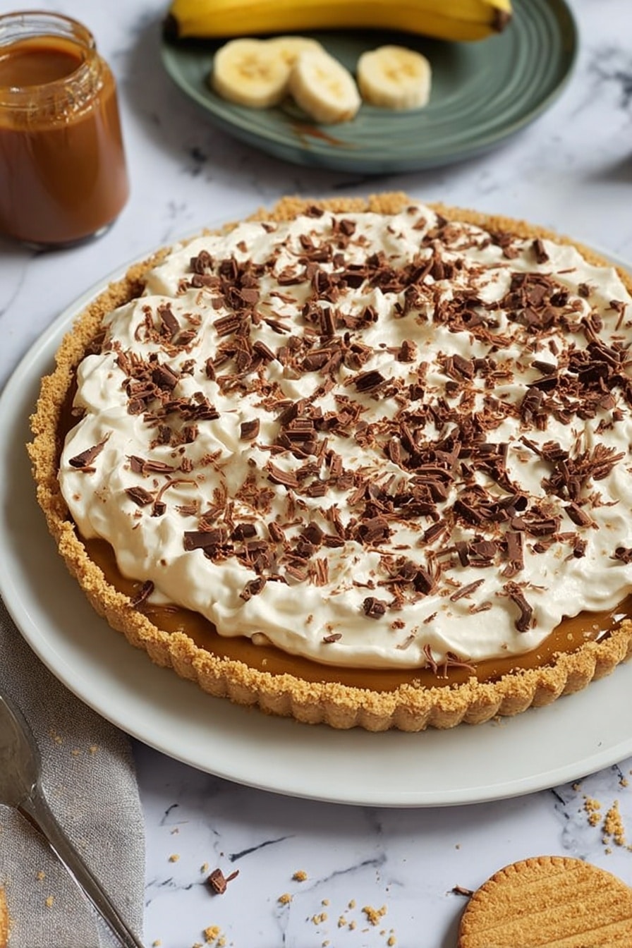 The image shows a dessert tart on a white plate with a crumbly golden brown crust forming the base, filled with a caramel-colored layer visible under a thick top layer of fluffy, white cream spread unevenly. The cream is generously covered with small, dark brown chocolate curls scattered across the surface. Around the plate, there is a jar of caramel sauce, a partially peeled banana, and a round biscuit with a bite taken out, all set on a white marbled surface. Photo taken with an iphone --ar 2:3 --v 7 - Easy No-Bake Banoffee Pie, no-bake dessert recipes, simple banana pie, quick indulgent desserts, no oven banana pie