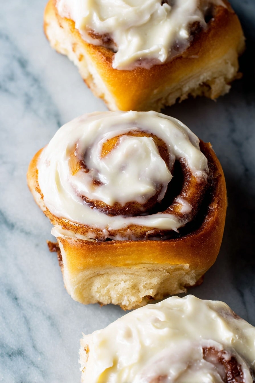 The image shows three cinnamon rolls with soft golden-brown spiral layers of dough swirled with cinnamon filling. Each roll is topped with a creamy white frosting that looks thick and smooth, spread unevenly with some parts thicker than others. The cinnamon rolls are placed on a white marbled surface that contrasts with their warm colors. The focus is close up, making the texture of the dough and frosting very clear. Photo taken with an iphone --ar 2:3 --v 7 - Ultimate Fluffy Cinnamon Rolls, homemade cinnamon rolls, soft cinnamon rolls with cream cheese frosting, easy cinnamon roll recipe, melt-in-your-mouth cinnamon rolls