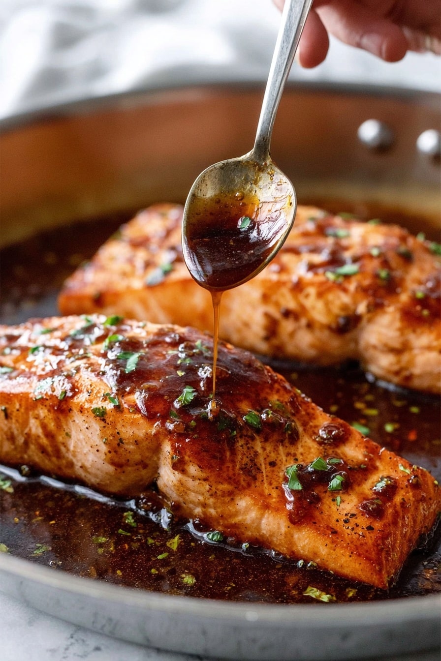 Two thick salmon fillets with a shiny dark brown sauce cover. The top layer is glossy, sticky sauce with small green herb bits sprinkled over. The salmon’s surface is textured with light char marks and a golden brown color under the sauce. A woman's hand holds a metal spoon above the front fillet, drizzling more sauce from the spoon onto the fish. The fillets sit in a shallow pool of the dark sauce within a metal pan. The background has a soft, white marbled texture. photo taken with an iphone --ar 2:3 --v 7 - Honey Garlic Glazed Salmon, Honey Garlic Salmon, Easy Salmon Recipes, Quick Salmon Dinner, Healthy Salmon Recipes