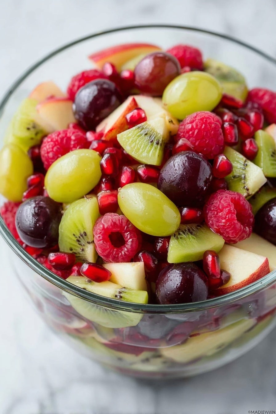 A clear glass bowl filled with a colorful mix of fresh fruits on a white marbled surface. The fruit salad has three main layers: the bottom layer is mixed with light green apple pieces with a smooth texture and thin red skin, the middle layer features bright green and dark purple grapes that are oval and shiny, and the top layer is scattered with vibrant red raspberries that have a soft, bumpy texture and small pomegranate seeds adding shiny ruby red dots. The bowl is shown from above, with a lime slice partially visible at the top edge of the image and some loose raspberries and grapes scattered around the bowl. Photo taken with an iphone --ar 2:3 --v 7 - Festive Christmas Fruit Salad, Christmas fruit salad, holiday fruit salad, easy Christmas salad, colorful holiday recipe