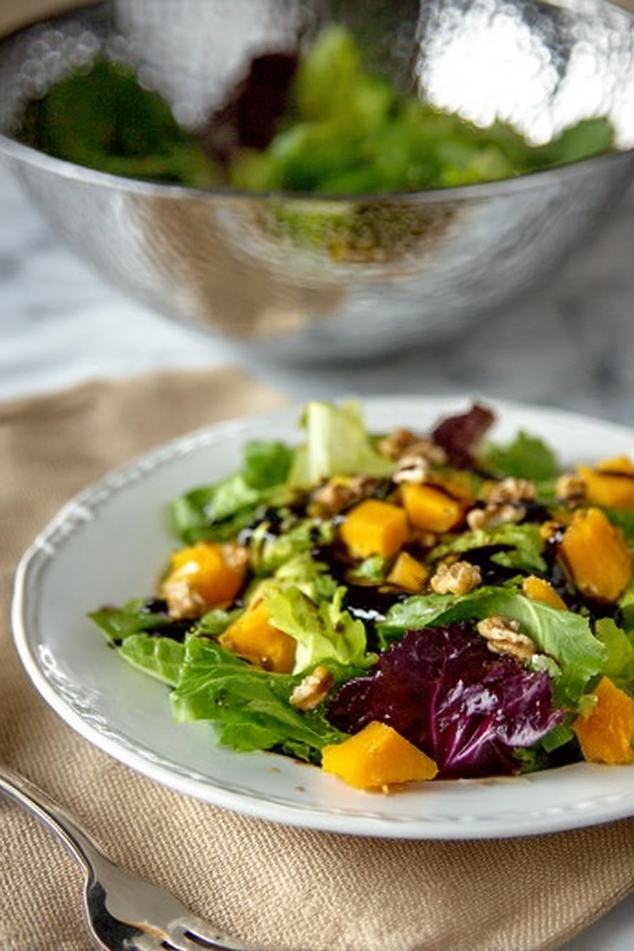 The image shows a white plate with a fresh salad on top placed on a beige cloth. The salad has several layers: a base layer of mixed green lettuce leaves that are bright green and purple with a soft texture, scattered orange chunks of roasted pumpkin or squash spread evenly over the greens, and small pieces of walnuts sprinkled throughout. Some dark balsamic dressing is drizzled over the salad, adding a glossy shine. In the background, a silver textured bowl with more greens inside sits on a white marbled surface, slightly blurred. A fork is visible next to the plate on the left side. photo taken with an iphone --ar 2:3 --v 7 - Butternut Squash Salad with Apple Cider Vinaigrette, fall salad recipes, healthy squash salads, roasted butternut squash recipes, seasonal salad ideas