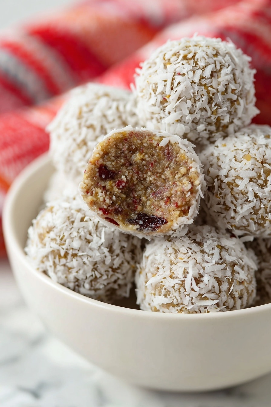 A close-up view of round dessert balls stacked inside a white bowl, each ball covered with a layer of white shredded coconut flakes, giving a rough texture on the outside. One ball is broken in half and shows a dense inside layer with a brown color mixed with visible tiny bits of red and light tan ingredients. The balls contrast against a soft white marbled surface in the background, with a red and white cloth slightly blurred behind the bowl. Photo taken with an iphone --ar 2:3 --v 7 - Healthy Date Energy Balls, natural snack bites, no bake energy balls, healthy snack ideas, quick healthy treats