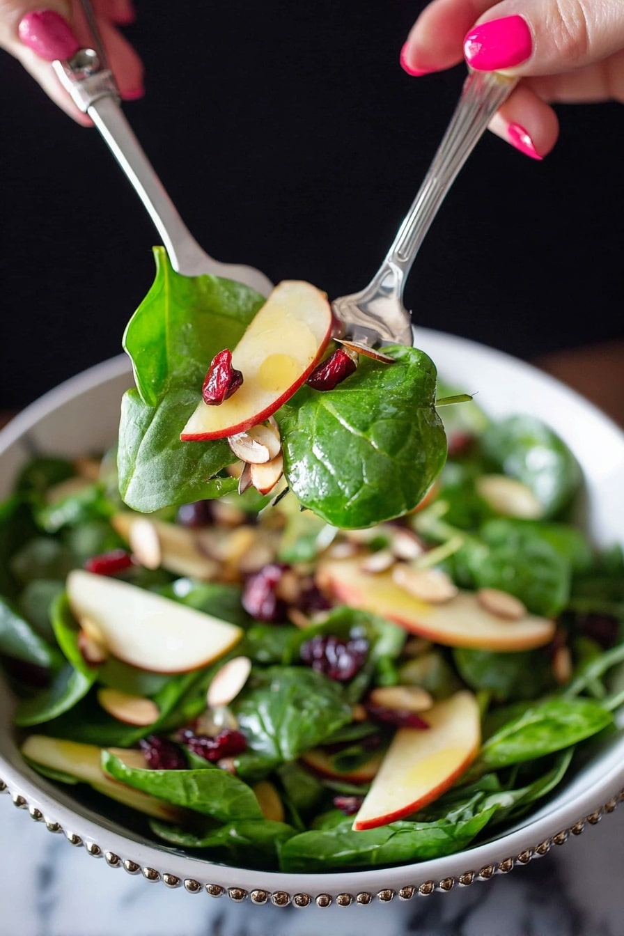 A close-up of a fresh salad being lifted by two silver utensils held by a woman's hand with pink nail polish. The salad has bright green spinach leaves, thin light yellow apple slices with red edges, small dark red dried cranberries, and light brown sliced almonds mixed inside a white bowl with a beaded rim. The background is dark, contrasting with the vibrant colors of the salad, and the surface underneath the bowl is a white marbled texture. photo taken with an iphone --ar 2:3 --v 7 - Apple Cranberry Spinach Salad, healthy fruit and vegetable salad, festive holiday salad, easy seasonal salad, fresh spinach and berry salad