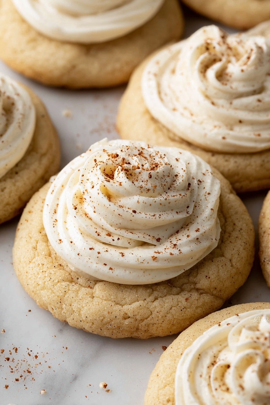 The image shows several round cookies with a light beige color, each having a thick base layer that looks soft and slightly crumbly. On top of each cookie is a swirl of white cream frosting, piped neatly in a circular shape with a peak in the center. Light brown spice sprinkles are scattered on top of the cream, adding texture and detail. The cookies are stacked close to each other on a white marbled surface. photo taken with an iphone --ar 2:3 --v 7 - Eggnog Thumbprint Cookies, holiday eggnog cookies, festive thumbprint cookies, Christmas baking ideas, boozy eggnog cookies