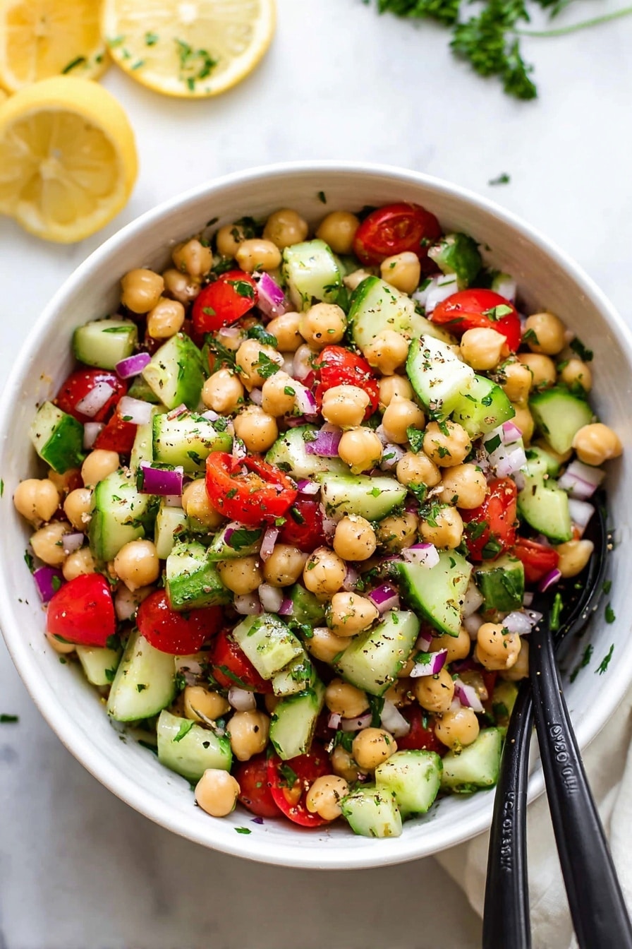 A white bowl filled with a fresh salad showing about four visible layers: the bottom layer is bright beige chickpeas, the next layer is made of medium-sized green cucumber chunks with darker green skin, followed by red tomato pieces, and small chopped purple onion mixed throughout. The salad is sprinkled with small bits of green herbs and black pepper. Two black serving spoons rest inside the bowl, angled toward the front. The bowl sits on a white marbled surface with some lemon halves and green parsley blurred in the background photo taken with an iphone --ar 2:3 --v 7 - Refreshing Chickpea Salad with Cucumber and Tomato, healthy chickpea salad, quick vegetarian salad, vibrant summer salad, easy chickpea salad