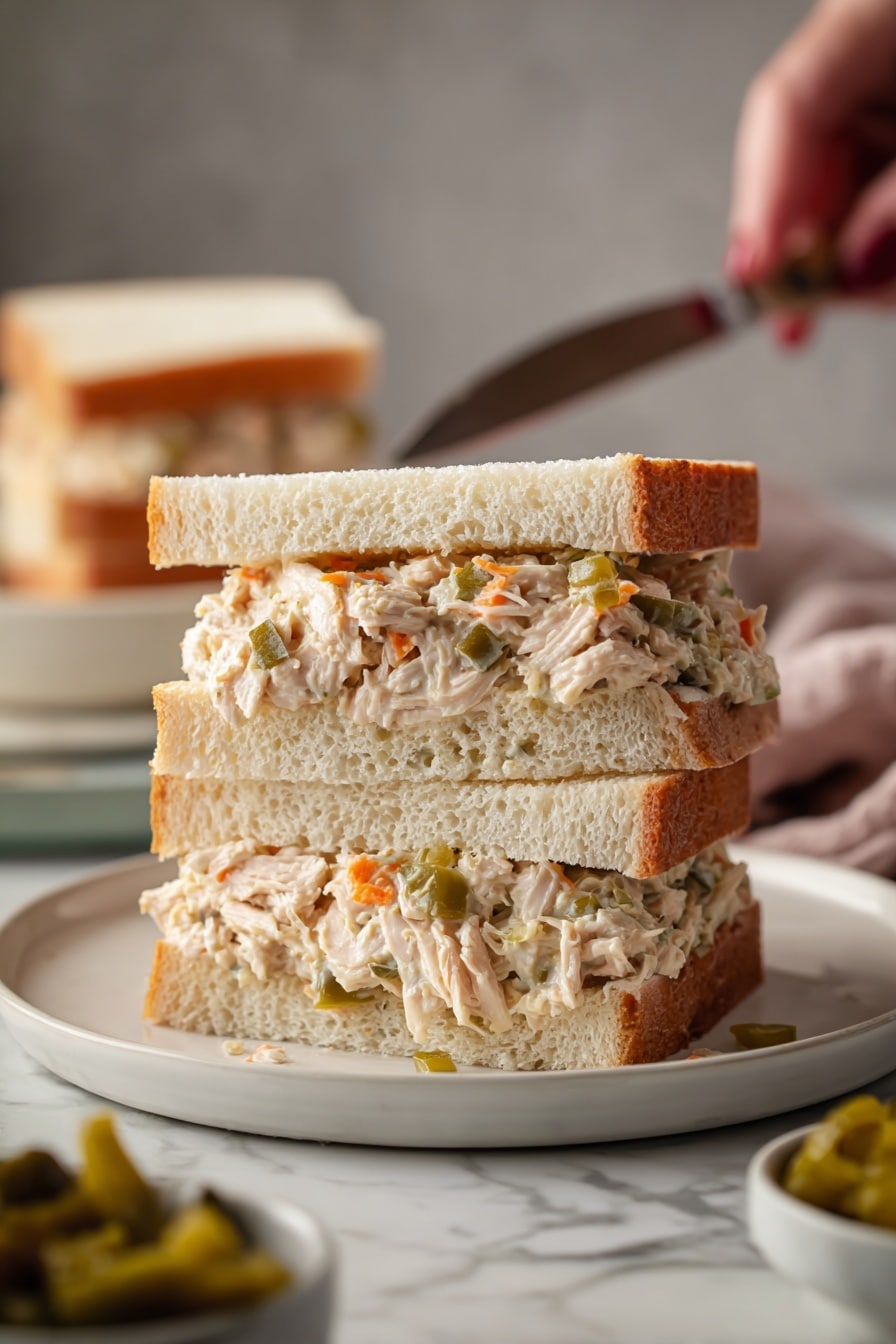 The image shows a sandwich cut in half and stacked on a white round plate. The sandwich has three layers from bottom to top: soft white sandwich bread with a fluffy texture, a thick layer of shredded cooked chicken mixed with small pieces of orange and green pickled vegetables, and another piece of white sandwich bread on top. Some filling spills out slightly from the sides, showing the moist and tender texture of the chicken and vegetables. In the background, more sandwiches and a small bowl with pickled vegetables can be seen on a white marbled surface. Woman's hand holding a knife is visible near the plate. The overall scene is warm and inviting, with soft lighting. photo taken with an iphone --ar 2:3 --v 7 - Slow Cooker Italian Chicken Sandwiches, Italian chicken sandwiches, easy slow cooker chicken recipes, flavorful chicken sandwiches, game day chicken sandwiches