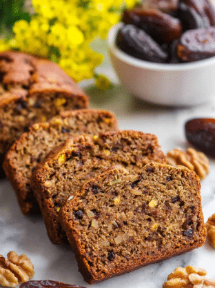 A close-up of four slices of brown nut bread lined up diagonally on a white marbled surface. Each slice shows a dense texture with visible dark brown nuts and darker bits inside the bread. Around the slices are loose whole walnuts and dark brown dried dates scattered. In the background, there is a white bowl filled with more dried dates and a soft-focused cluster of yellow flowers. The scene is bright and natural. photo taken with an iphone --ar 2:3 --v 7 - Date Nut Bread, moist date nut bread, healthy quick bread, homemade date bread, easy date nut bread recipe